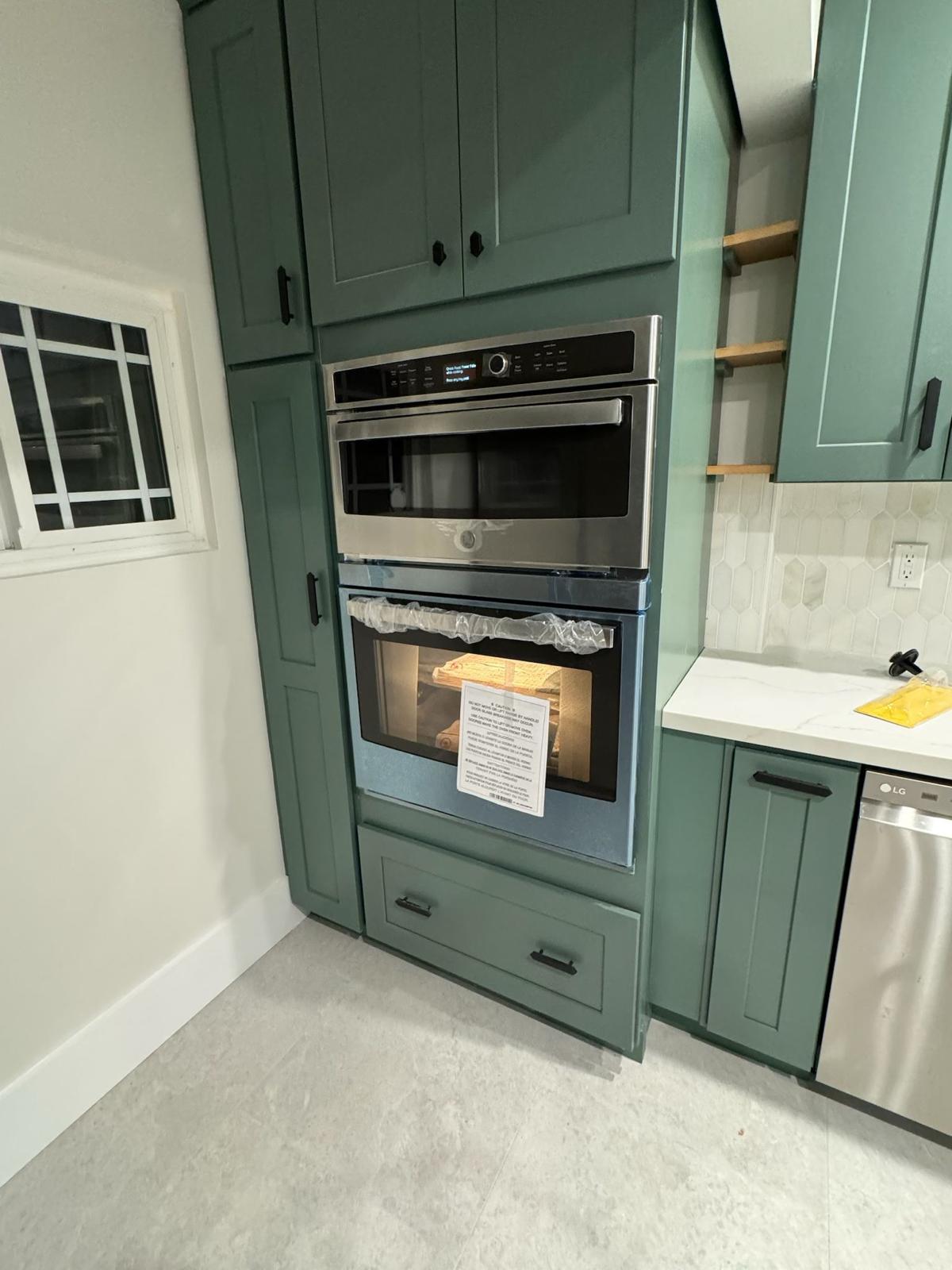 Built-in wall oven and microwave column flanked by green cabinetry and open oak shelving